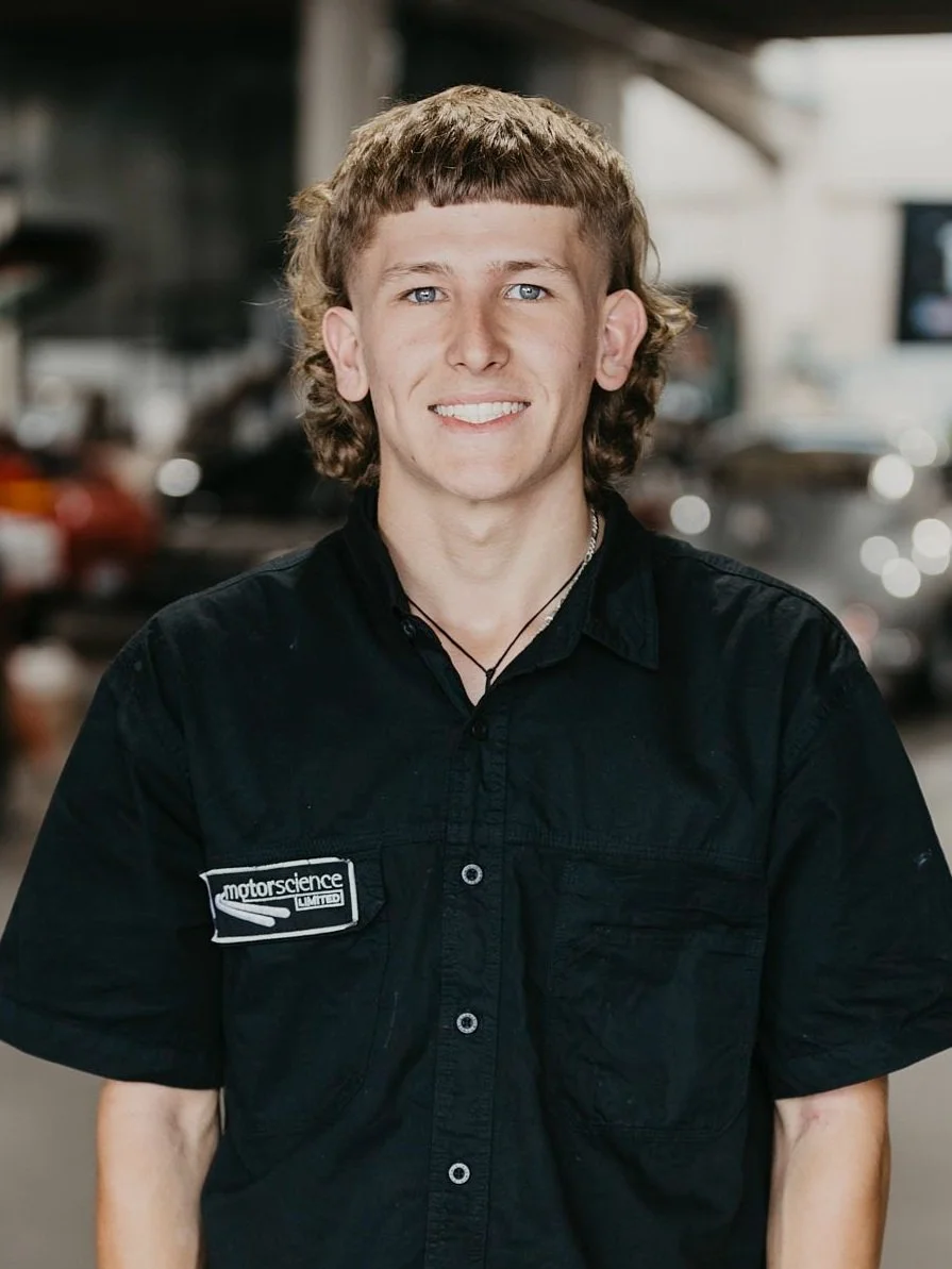 Young man with light skin, curly light brown hair, blue eyes, wearing a black shirt with a motorsport logo, smiling, standing in front of the workshop setting with Porsche cars in the background.