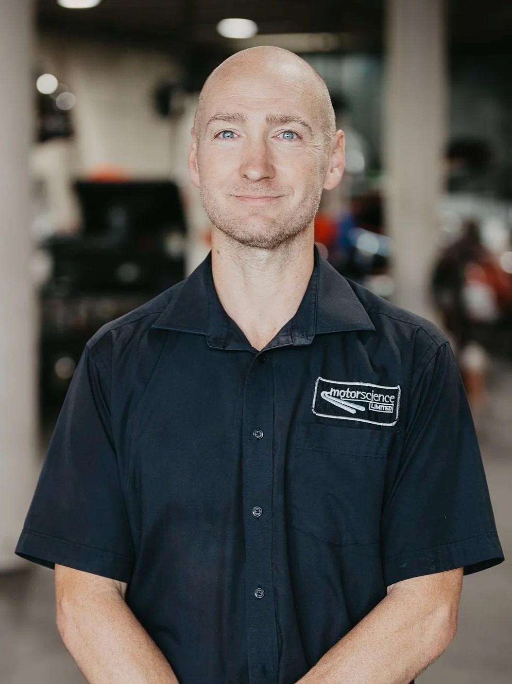 A man with a shaved head and blue eyes, wearing a dark navy polo shirt with a Motorsport Science Limited logo, standing in front of the workshop setting with Porsche cars in the background.