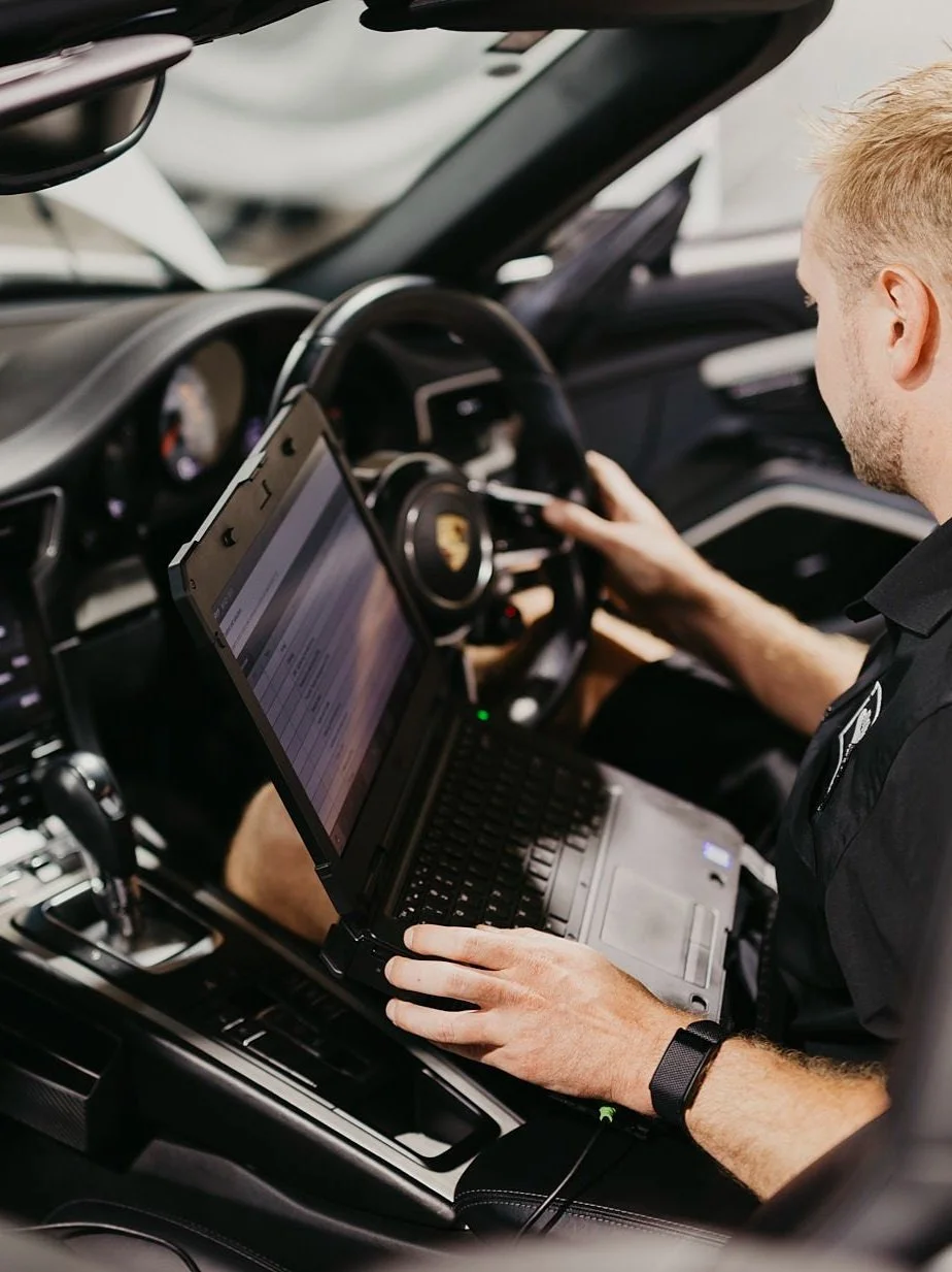A male technician in a black Motorscience uniform sitting inside a car, working on a diagnostic tool similar to a laptop, with the Porsche's steering wheel visible in front of him.