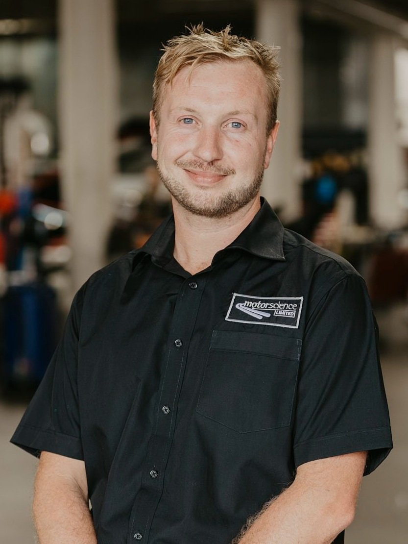 A Motorscience team member, Max Booth, smiling with blonde hair and a beard, wearing a black shirt with a 'motorscience' logo, standing in front of the workshop setting with Porsche cars in the background.