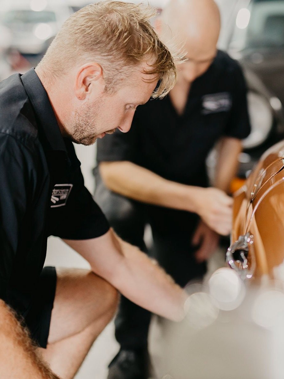 Two male technicians inspecting a tan Porsche car in the Motorscience garage, with one man in the foreground focusing on the vehicle and the other in the background.