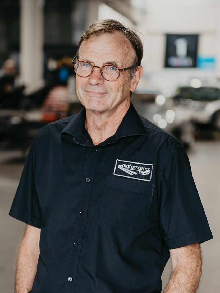 Motorscience Founder, Peter Booth, a middle-aged man with glasses, wearing a black collared shirt with a logo for 'Motorscience' stands in front of the workshop setting with Porsche cars in the background.