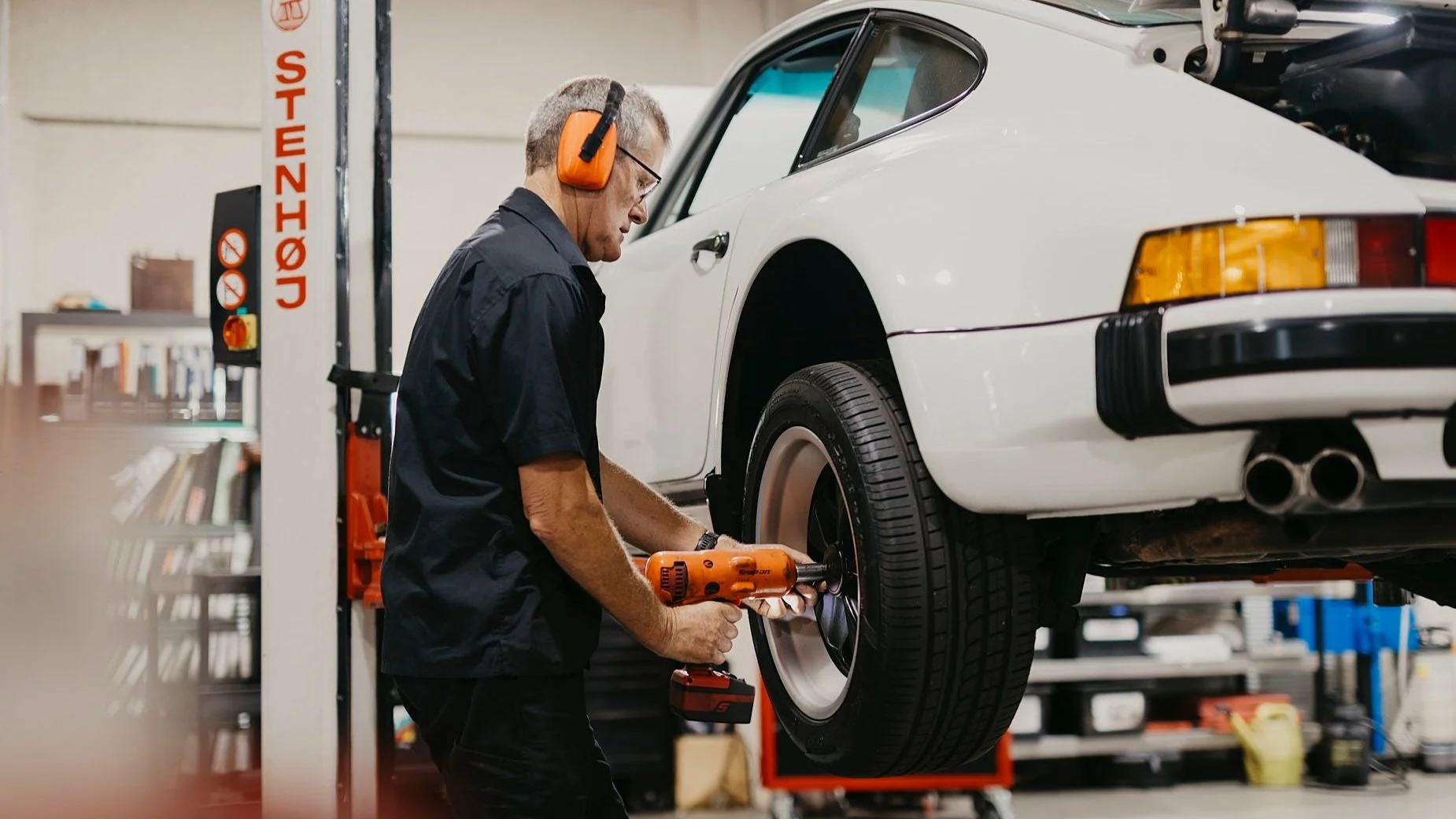 A male technician with grey hair wearing glasses, orange ear protection, and a black Motorscience shirt, working in an auto repair shop. He is using an orange power drill on a white Porsche sports car raised on a lift, focusing on the wheel.