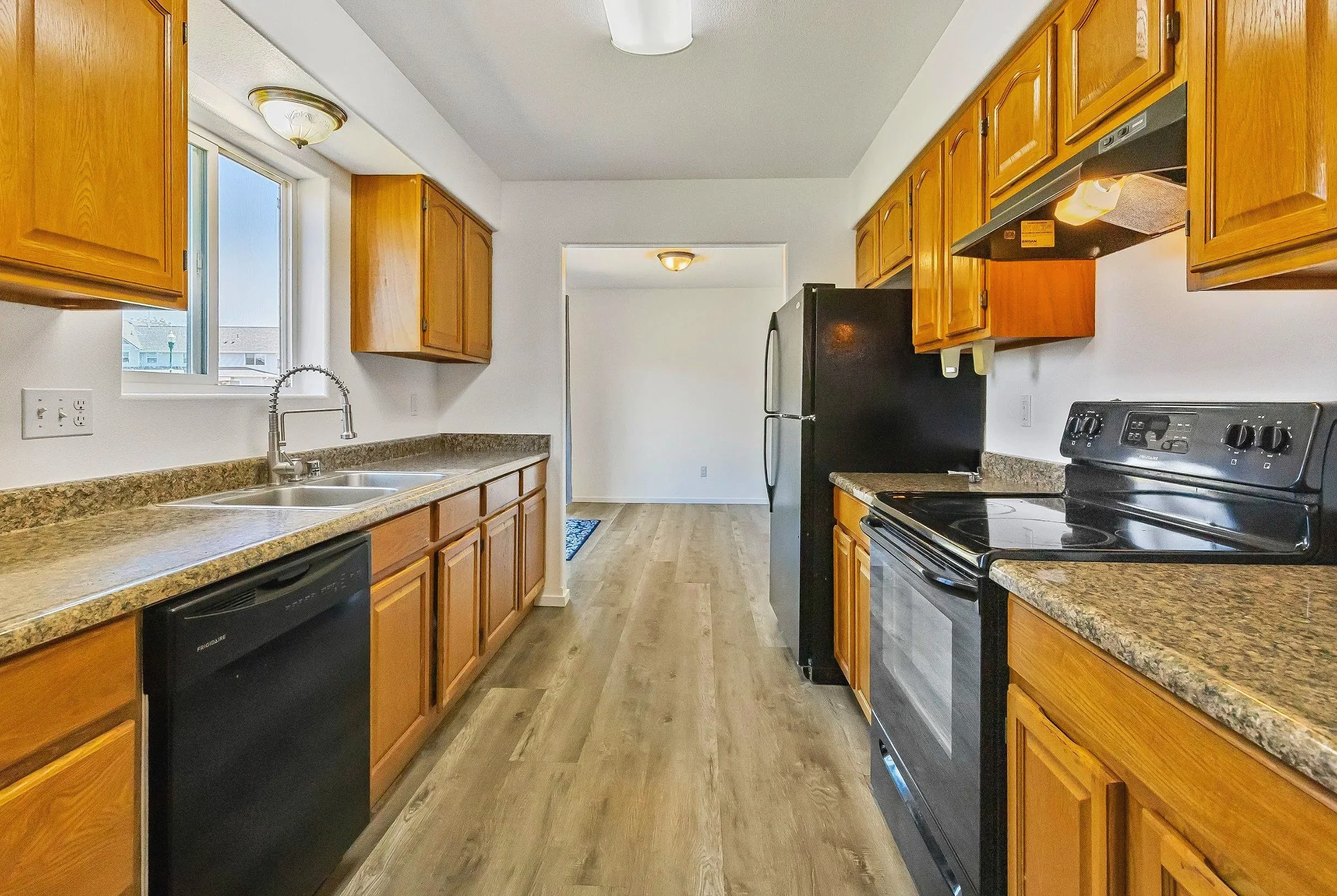 Kitchen with wooden cabinets, black appliances, granite countertops, a window over the sink, and wood-look flooring.