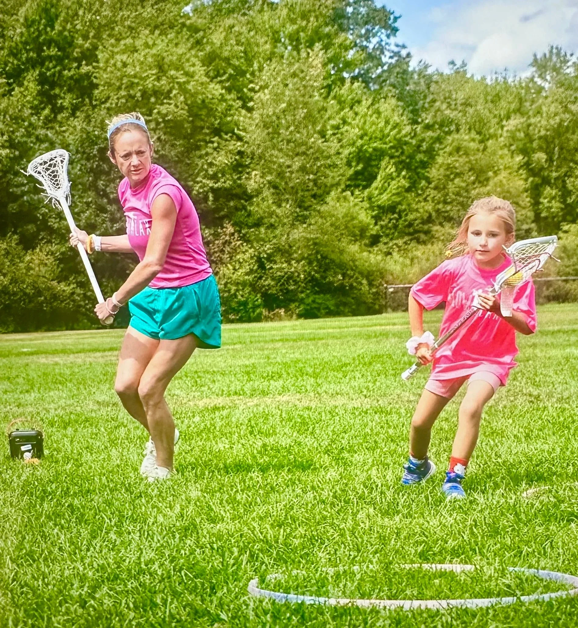 A woman and a girl playing lacrosse outdoors on a grassy field with trees in the background. The woman is in a pink shirt and teal shorts, holding a lacrosse stick, looking to her left. The girl is in a pink shirt and pink shorts, running with a lacrosse stick in her right hand, and appears focused.