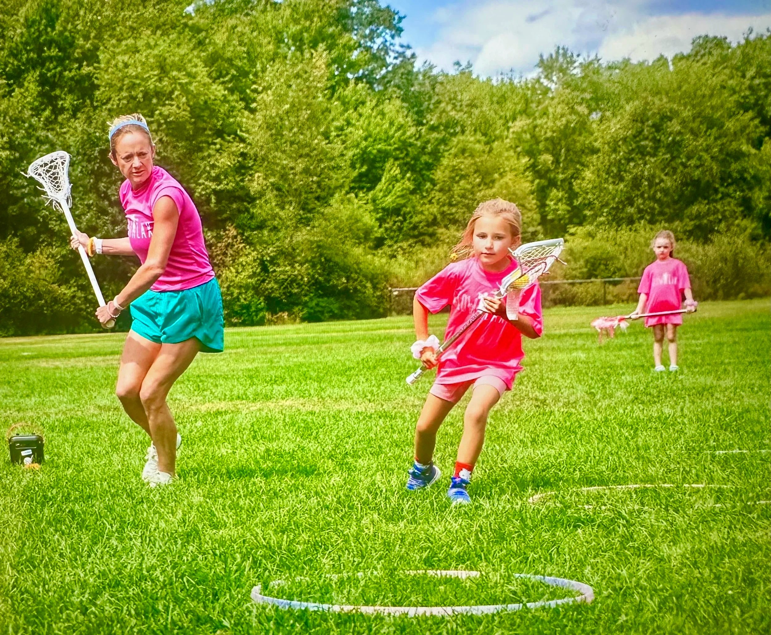 Three girls playing lacrosse outdoors on a grassy field with trees in the background. The girl in the foreground is running and holding a lacrosse stick. The other two girls are in the background, also holding lacrosse sticks and running.