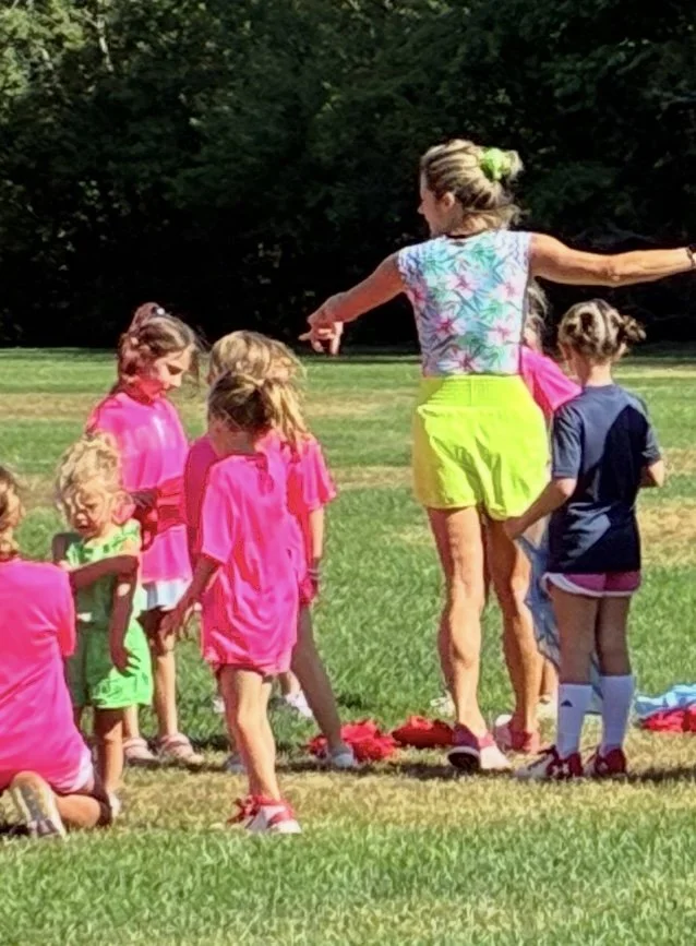 A group of children and an adult woman standing outdoors on a grassy field, possibly during a sports practice or game, with trees in the background.