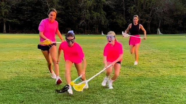Four women wearing pink and black athletic clothing playing lacrosse on a grassy field with trees in the background.