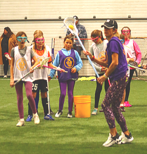 Children participating in an indoor team-building activity with a woman guiding them, holding pool noodles, in a sports facility.