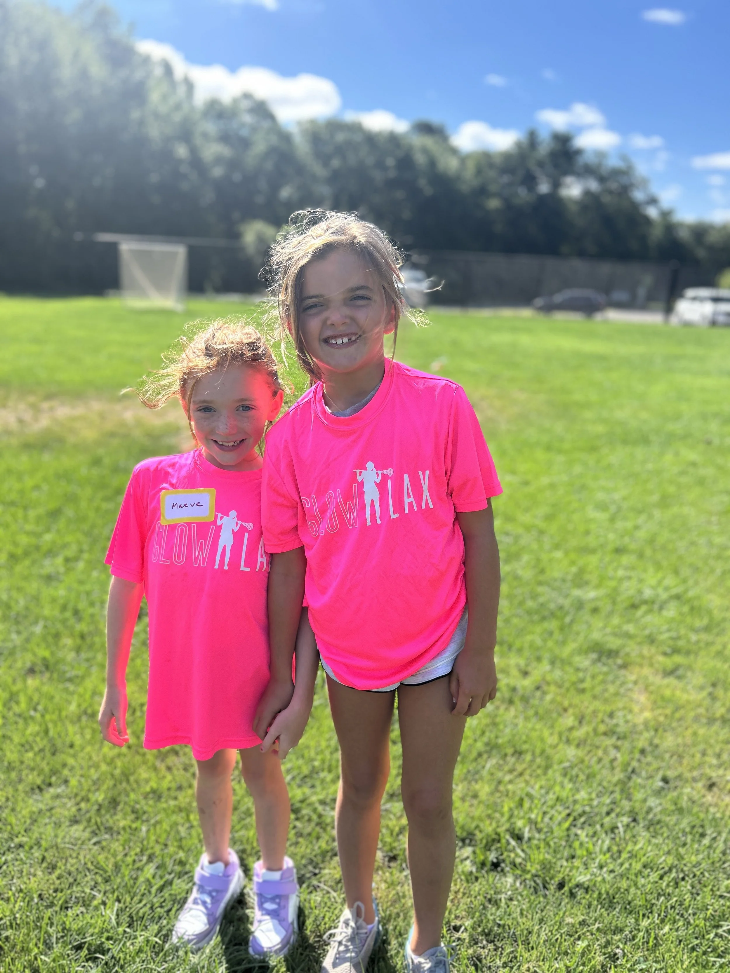 Two young girls standing on a grassy field, smiling at the camera, wearing bright pink shirts that say "Glow & Relax," with trees, a clear blue sky, and a fence in the background.