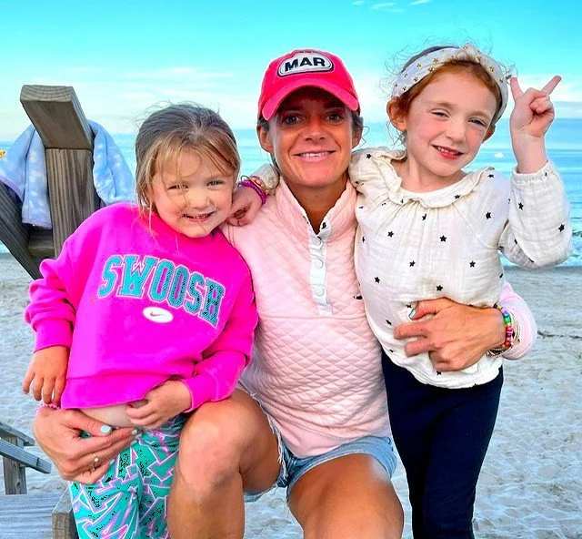 A woman and two young girls smiling on a beach, with the woman in a pink cap and pink attire, and the girls in colorful sweaters, one pink and one white, embracing each other.