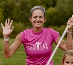 Coach Caroline smiling outdoors, waving with her right hand, holding a stick or pole with her left hand, wearing a pink athletic shirt, in a grassy area with trees in the background.