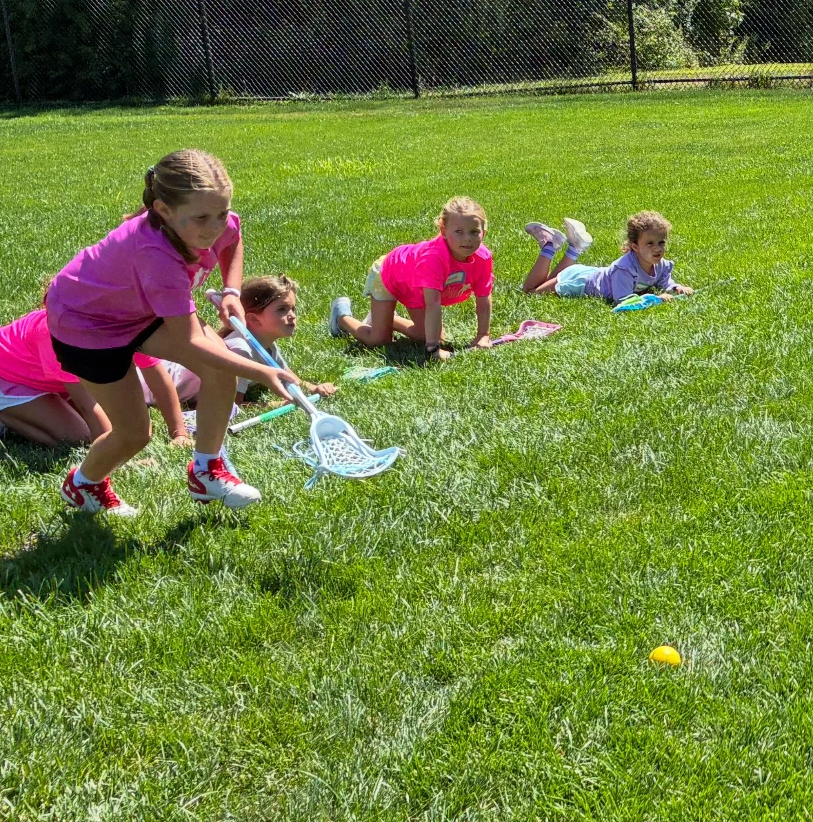 Four young girls in pink/t-shirts and shorts, crawling or lying on the grass during an outdoor activity, with a tennis ball nearby.