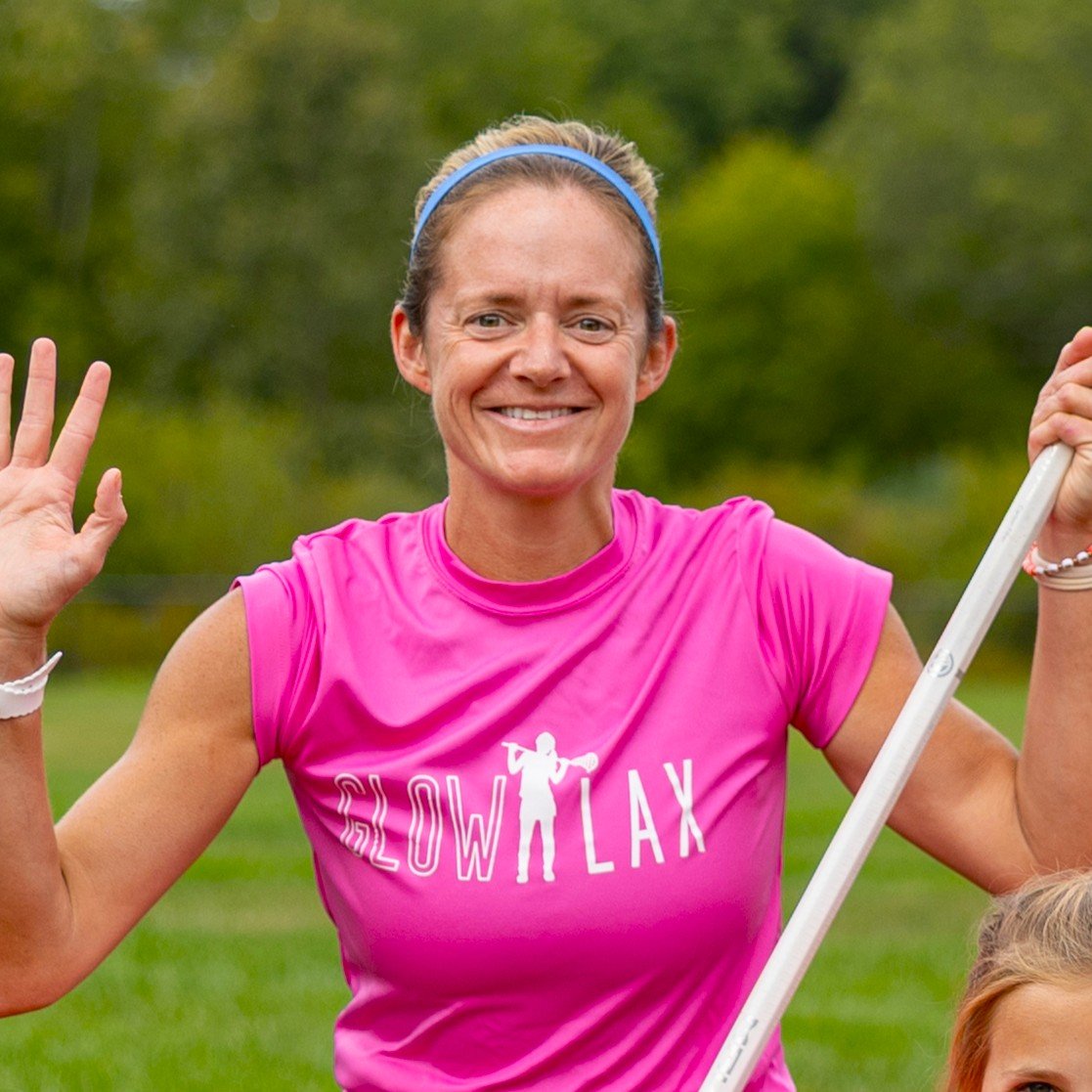 A smiling woman in a pink athletic shirt waving and holding a lacrosse stick outdoors.