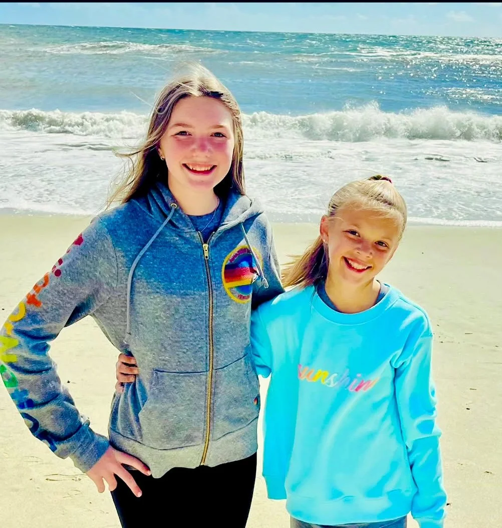 Two smiling girls standing on the beach with ocean waves and blue sky in the background.