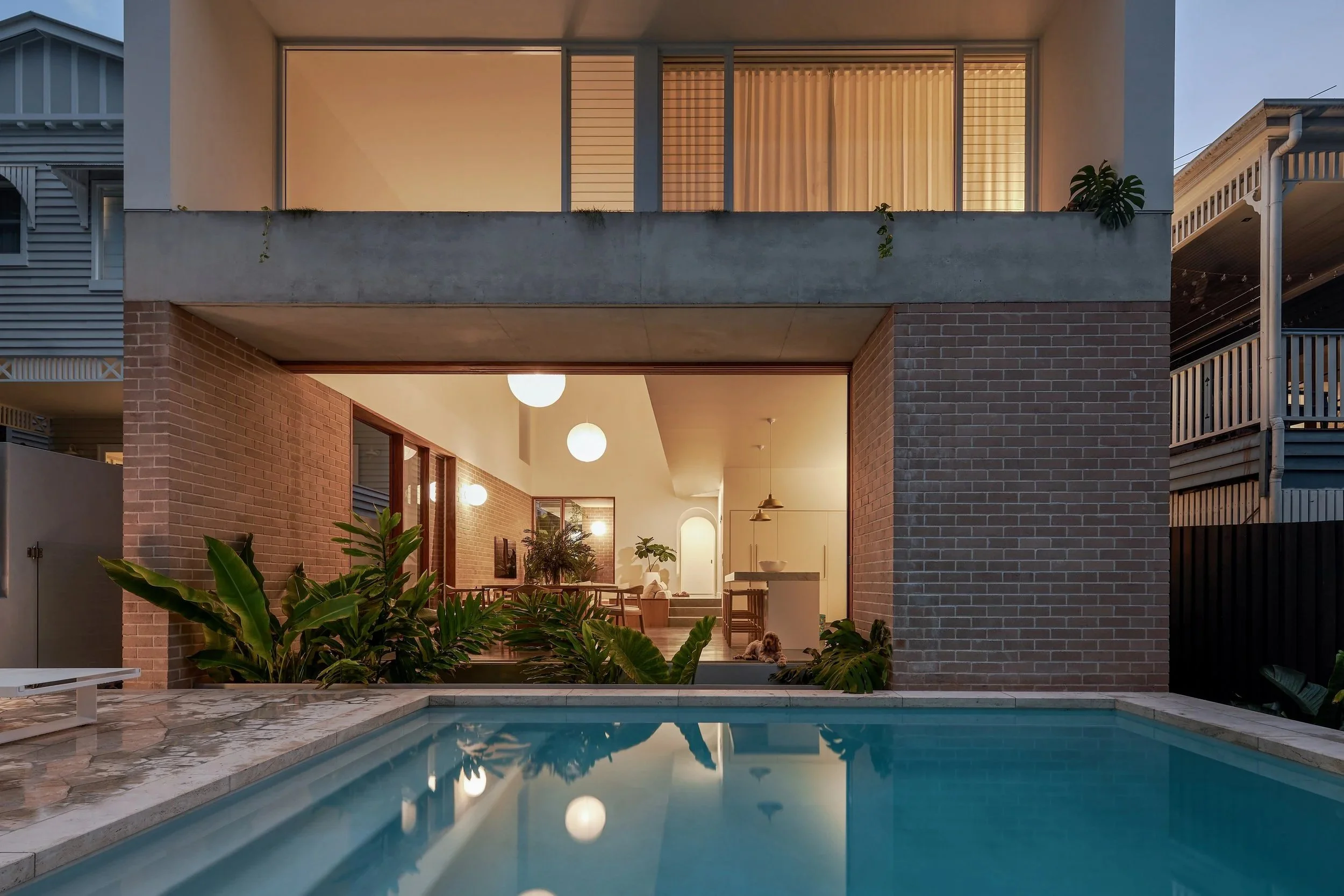 Modern house with brick and concrete exterior, large glass windows, and a backyard with a swimming pool, lush green plants, and a view into a well-lit living area.