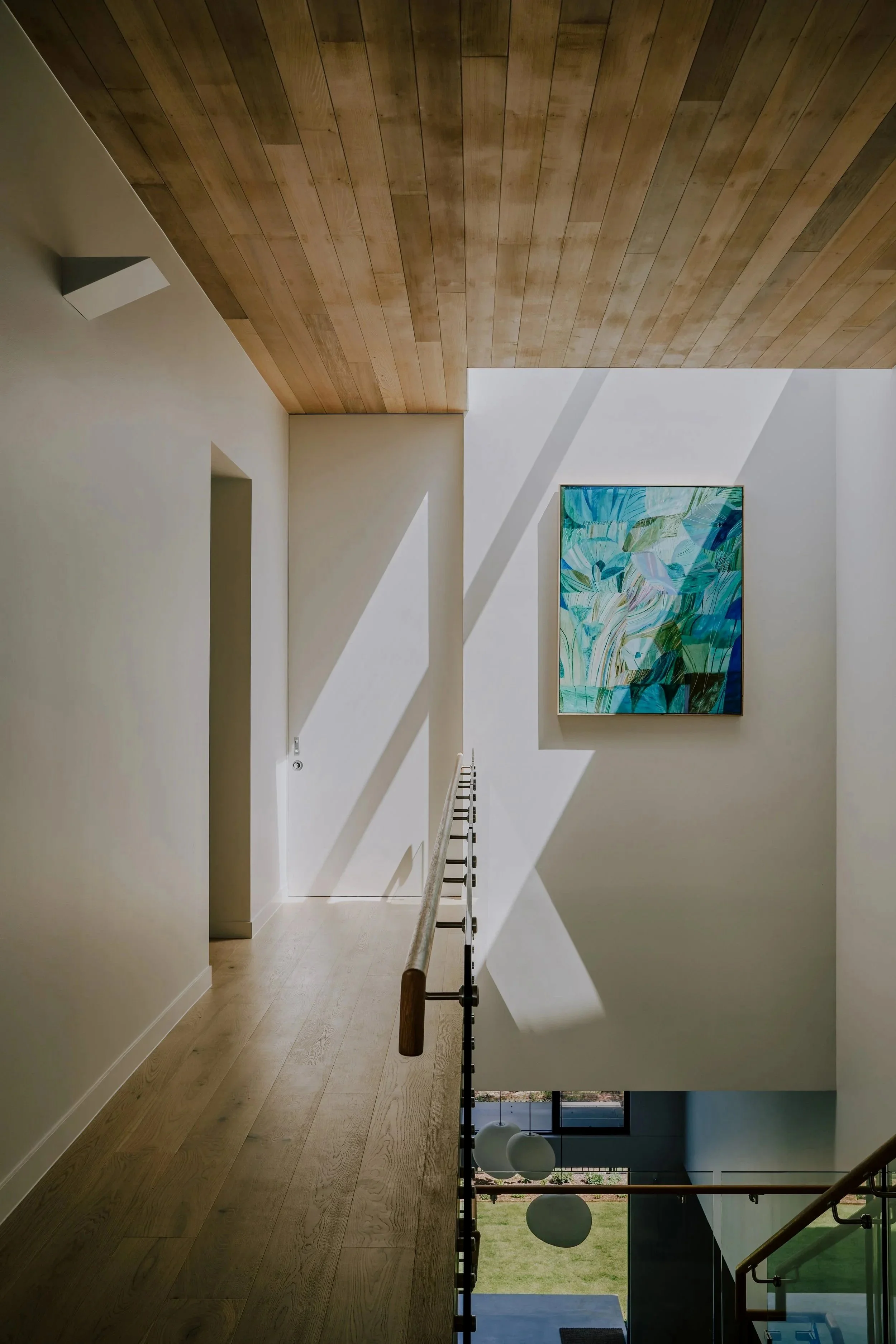 Interior view of a modern house with wooden ceiling, white walls, abstract blue and green painting, wooden handrail, and sunlight casting shadows.