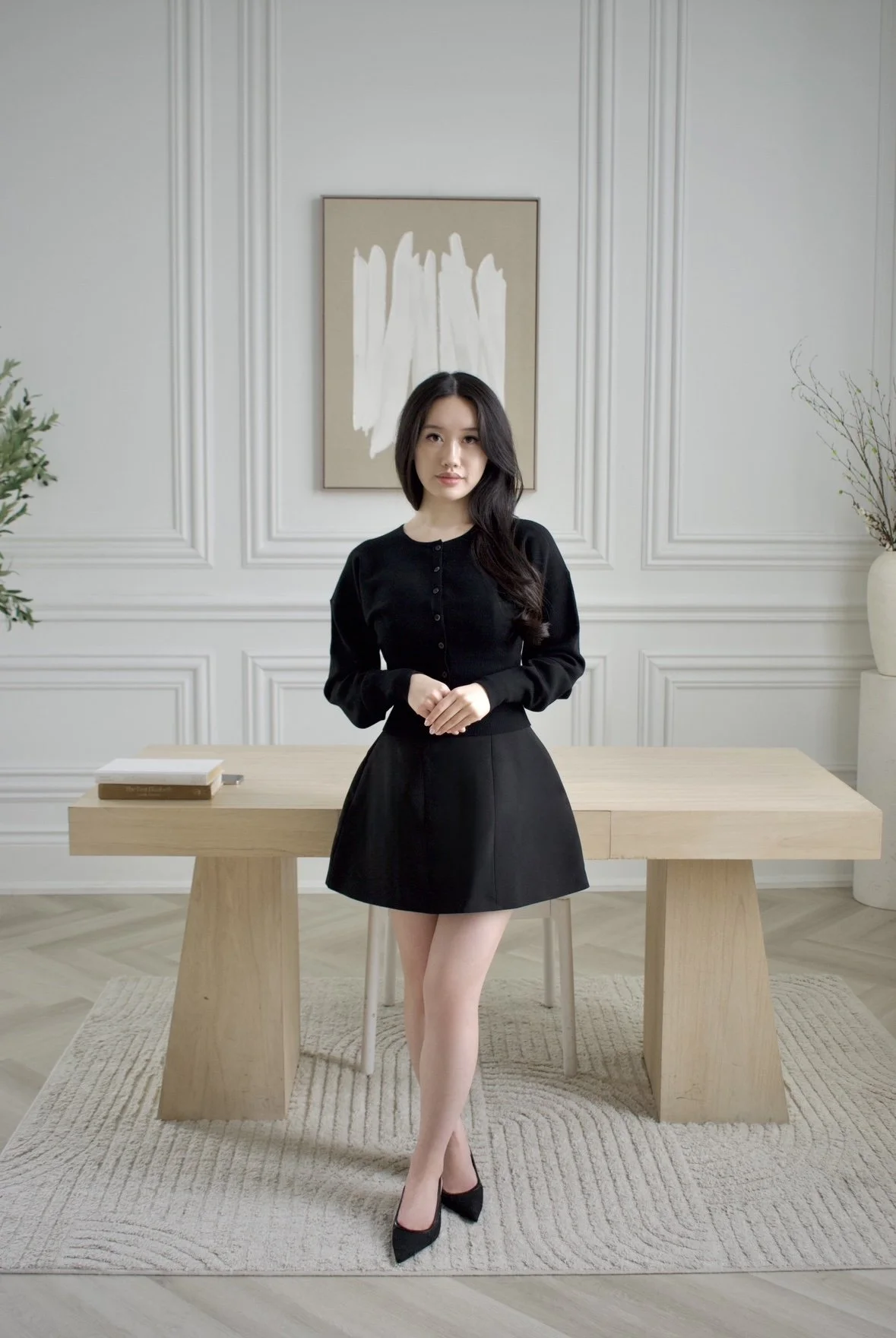 A young woman in a black long-sleeve top and black skirt standing in a modern, minimalist room with white walls, a wooden table, and abstract artwork hanging behind her.