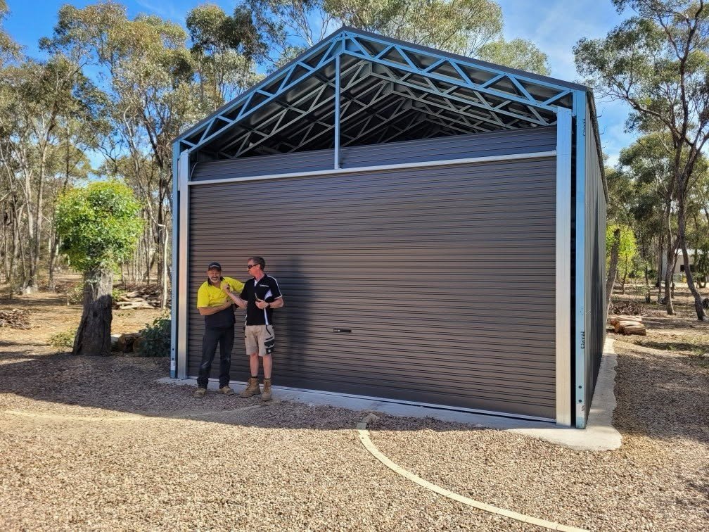 Two men standing in front of a large metal garage door, in an outdoor area with gravel ground and trees in the background.