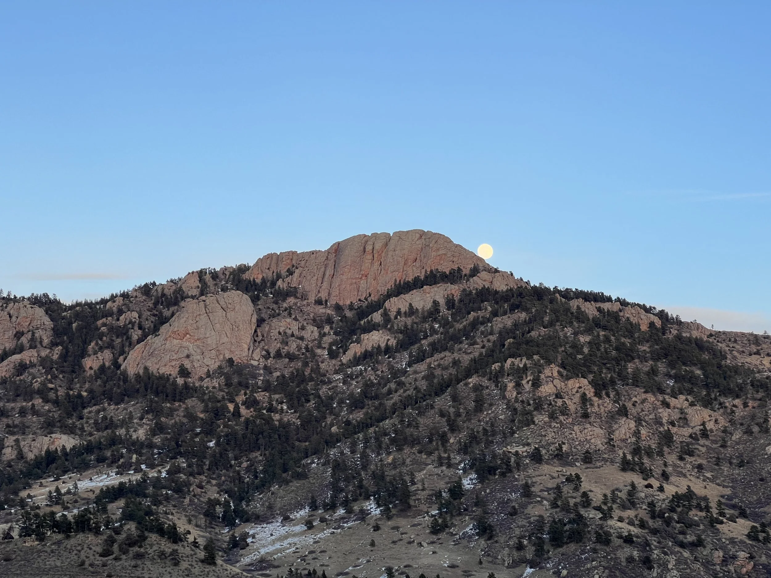 Mountain with rocky cliffs and sparse trees under a clear blue sky with the moon visible near the horizon.