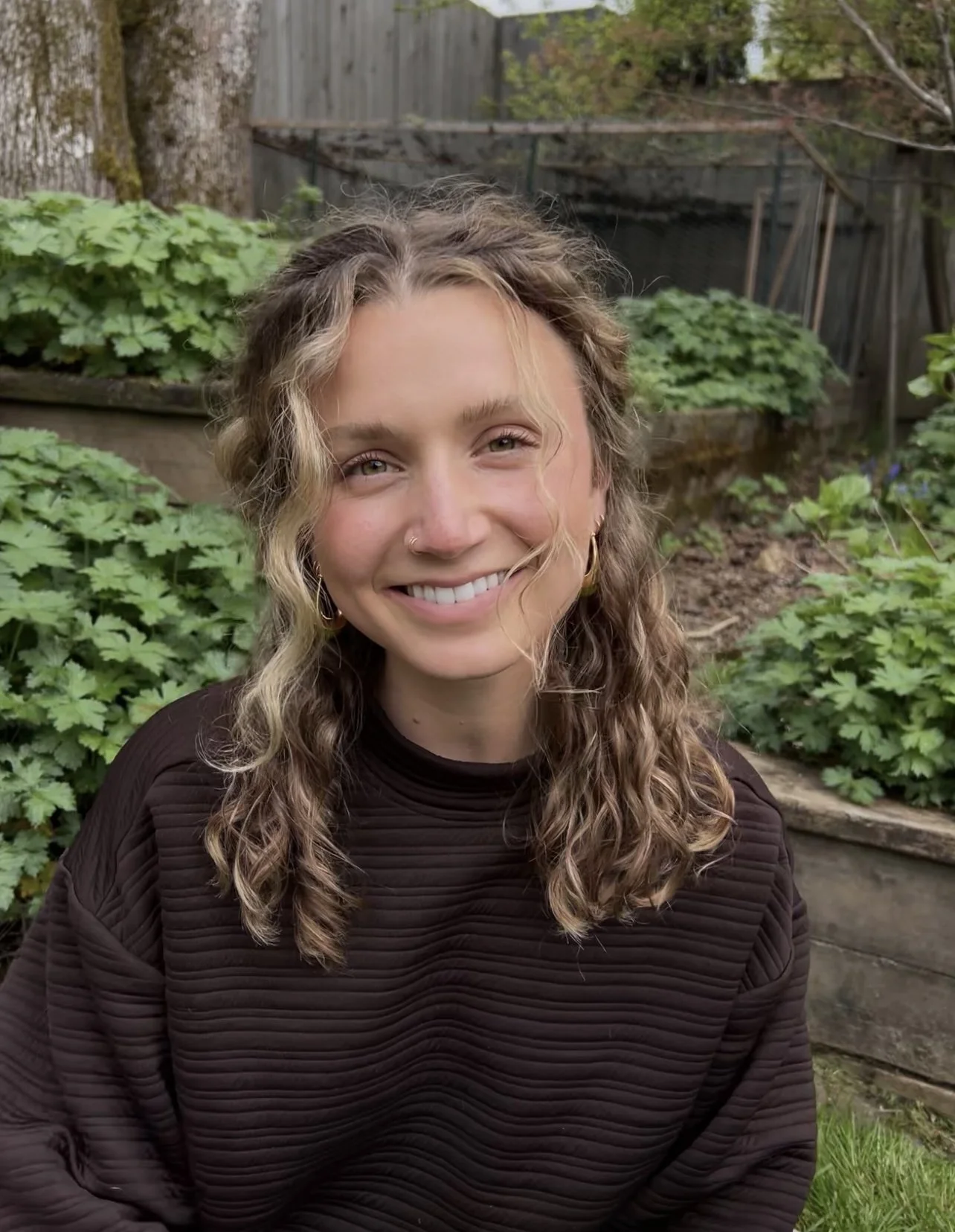 Woman with curly hair wearing a dark sweater, smiling in a garden setting with green plants and trees.