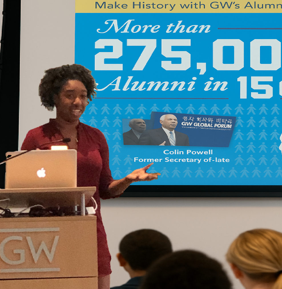 A woman is giving a presentation in front of a large screen that displays statistics about GW alumni, including more than 275,000 alumni in 150 countries, and features a photo of Colin Powell, a former Secretary of State. She is standing next to a ta