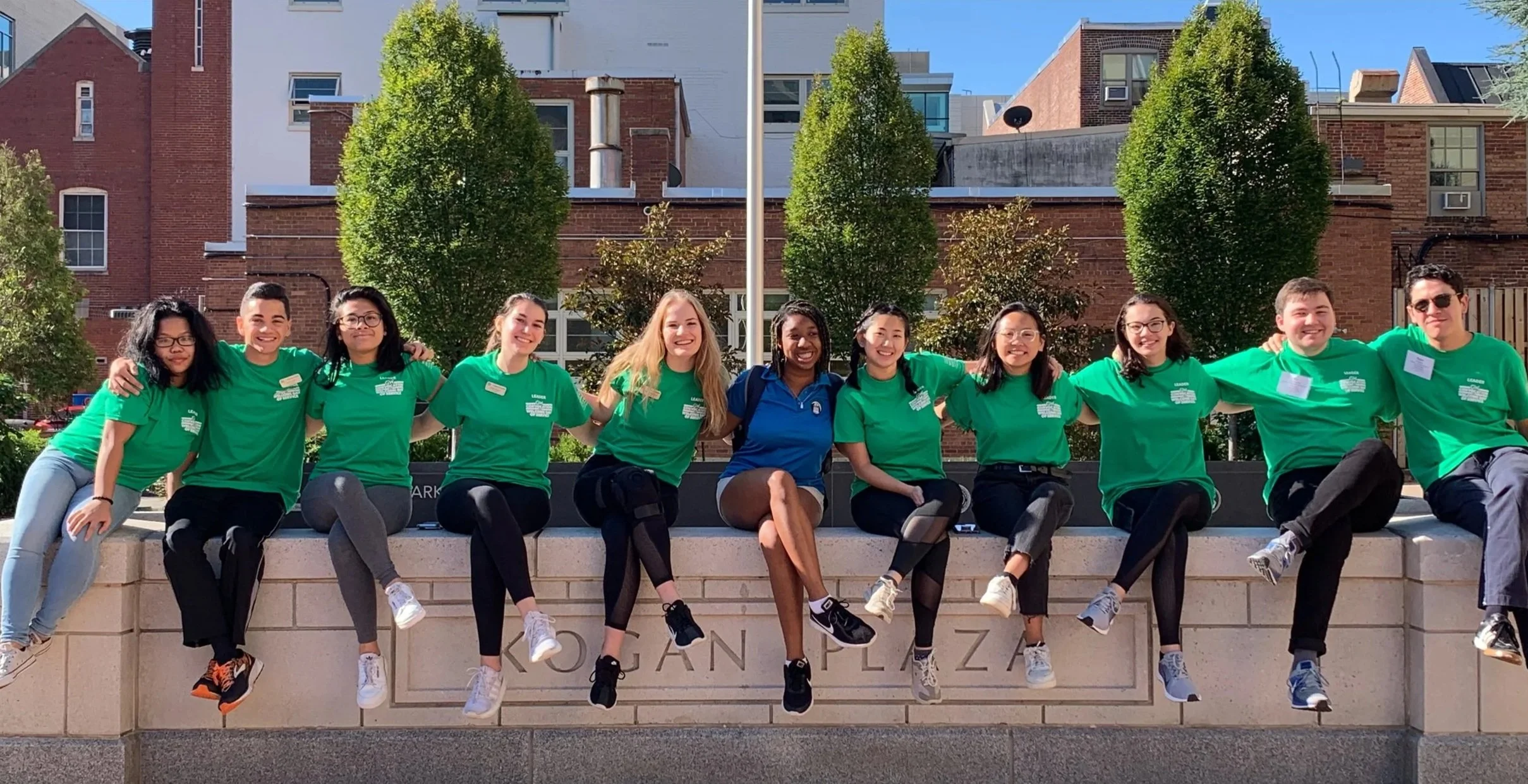 A group of ten diverse young adults sitting on a ledge outside, wearing matching green shirts, smiling with arms around each other, in front of a building with brick and white facades, trees, and a clear blue sky.