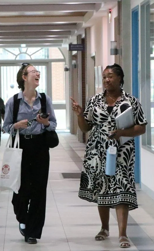 Two women walking and talking in a hallway, one young with glasses, the other older with braided hair, holding notebooks and a water bottle, smiling and gesturing.