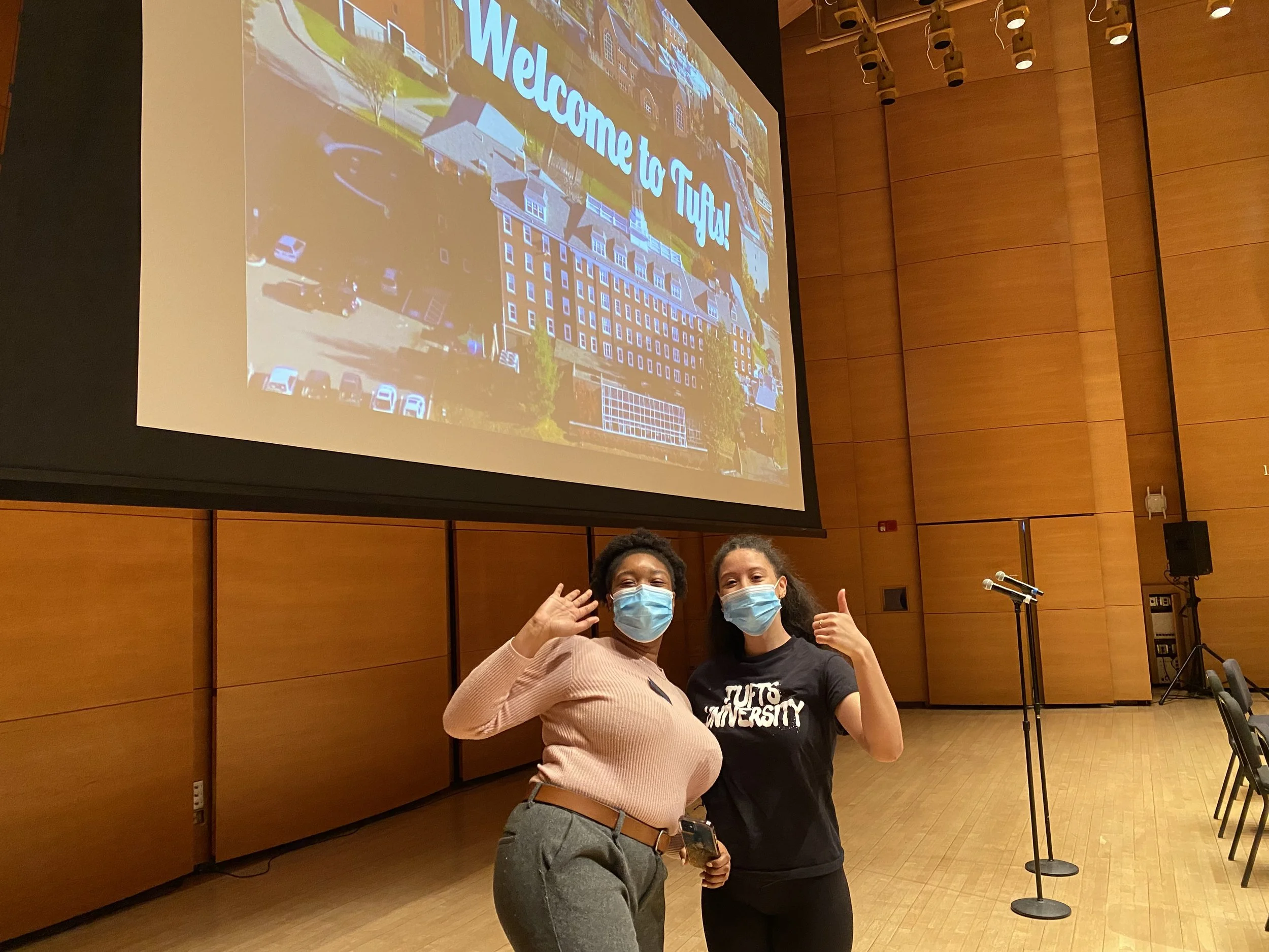 Two women standing in front of a large projected screen with a welcoming message. They are wearing masks, one with a pink sweater and gray pants, and the other with a black t-shirt with white text. The women are smiling and giving thumbs-up signs.