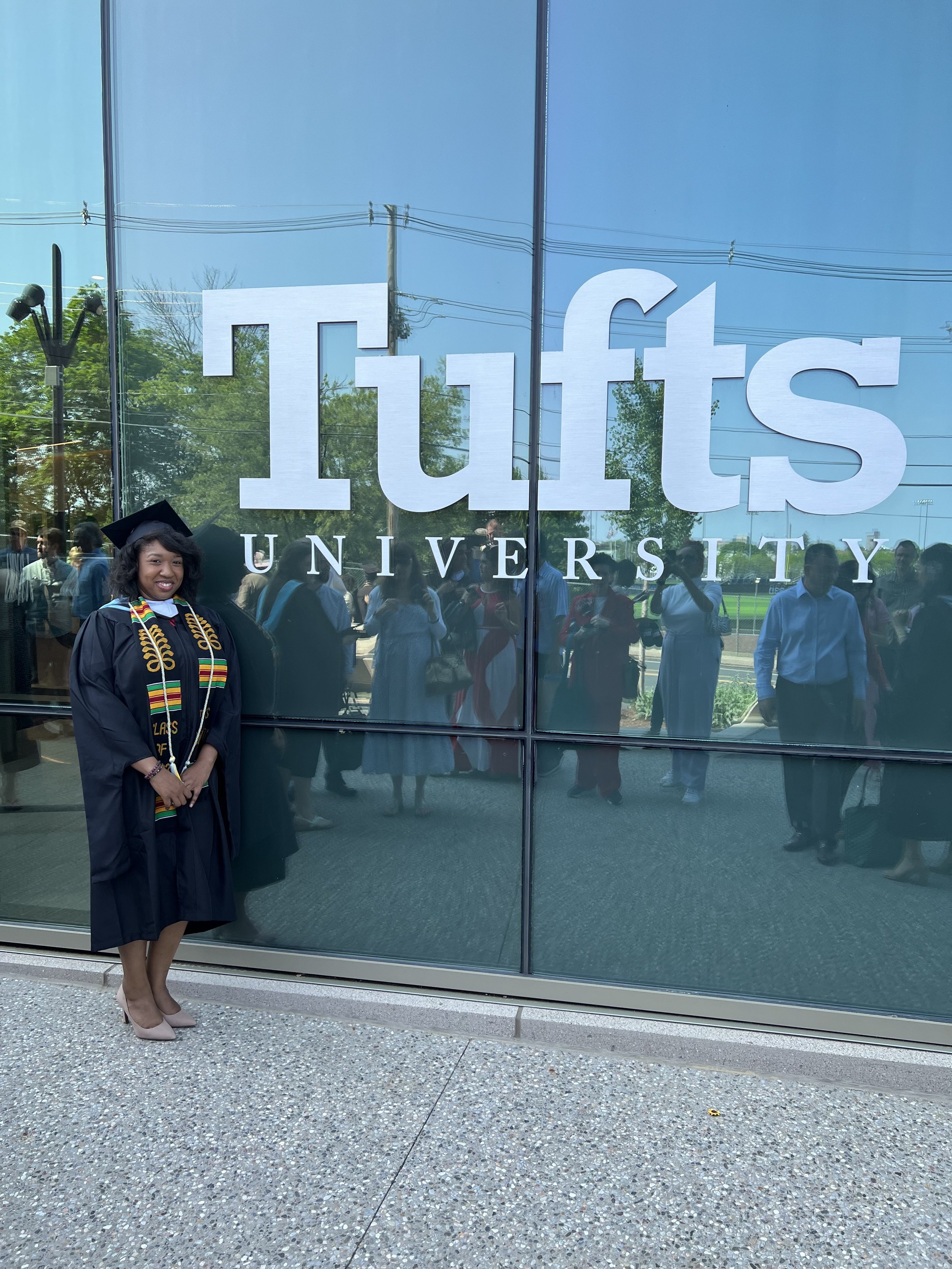 Woman in graduation cap and gown standing outside in front of a reflective glass wall with the words "Tulsa University" on it, during what appears to be a graduation ceremony.
