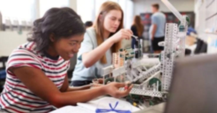 Two girls working together on a robotics project in a classroom or lab setting, assembling a white plastic and metal structure with various components.