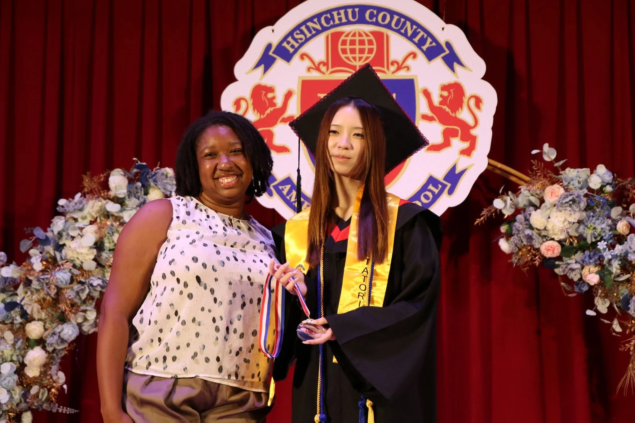 A young woman in graduation gown and cap standing on stage, shaking hands with a woman in a sleeveless polka dot blouse, in front of a red curtain and floral arrangements, with a school emblem in the background.