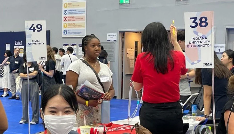 Students at a college fair, with signs for Miami University and Indiana University Bloomington, surrounded by people and booths.