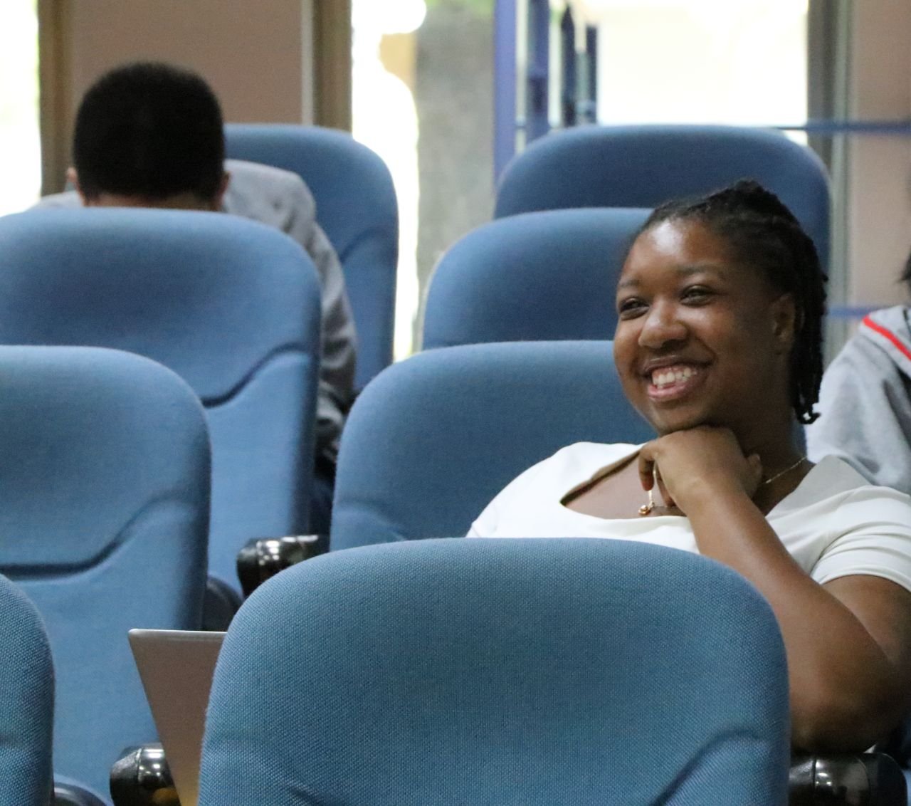 Smiling woman sitting in a blue seat on a bus or train, surrounded by empty blue seats, with a person in the background facing away.
