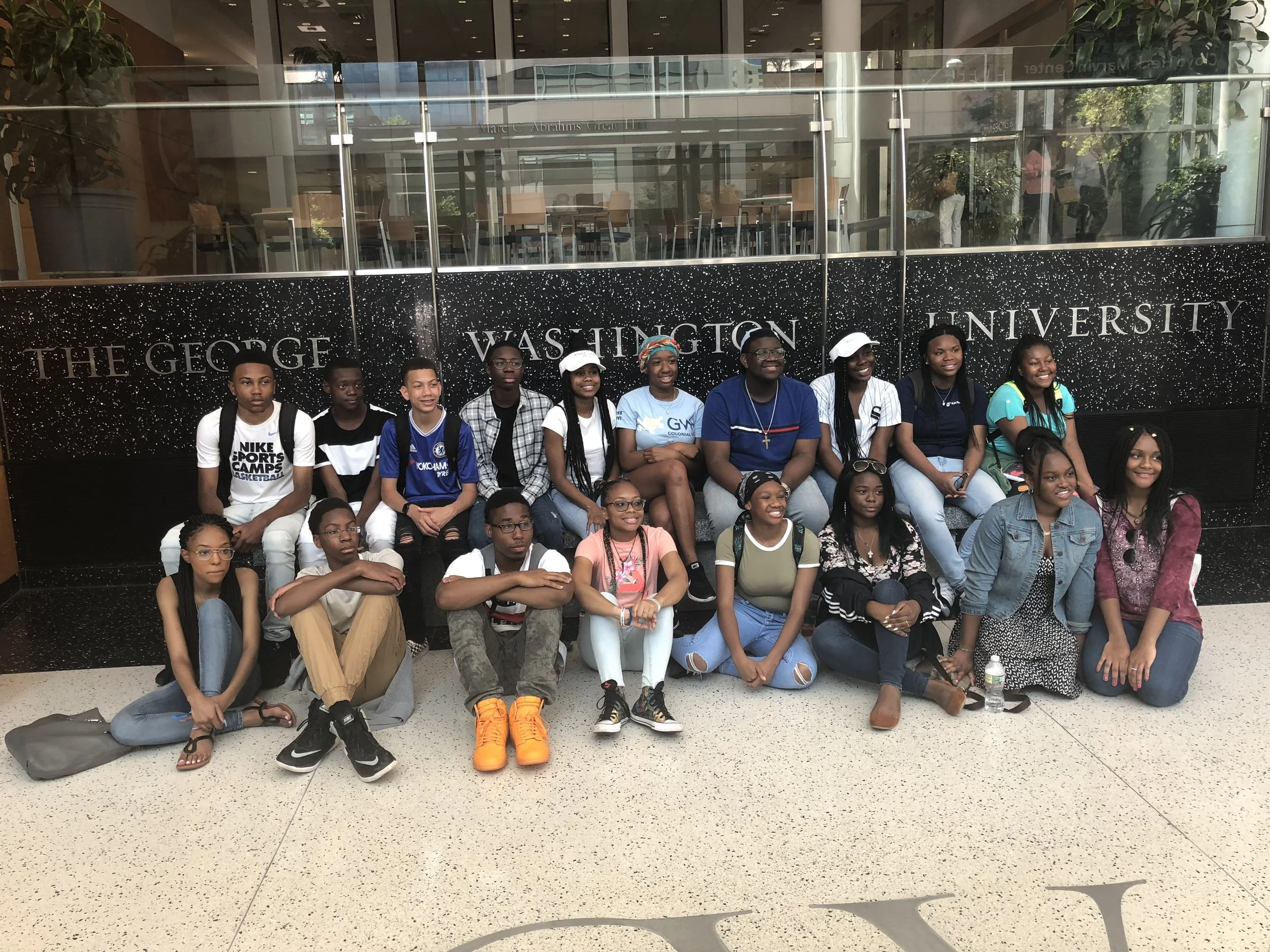 Group of diverse young people sitting on the floor in front of a sign that says 'The George Washington University' inside a building.