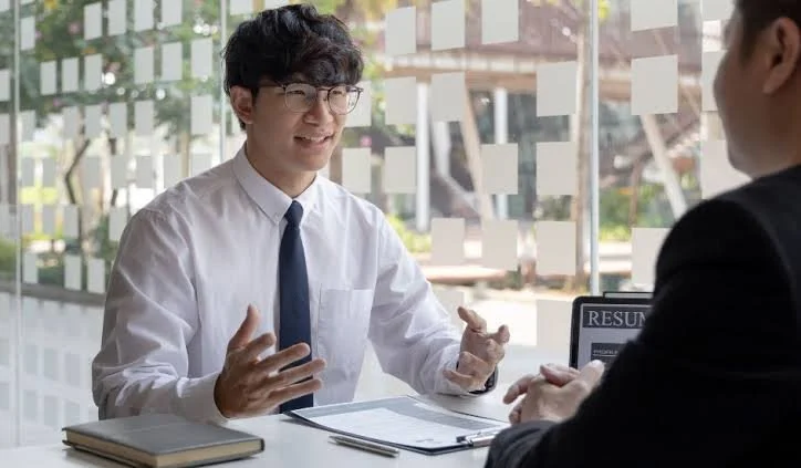 A job interview between a young man with glasses and a white shirt and a person in a suit, seated at a table in a glass-walled room.