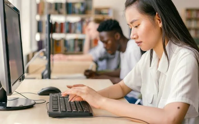 Young woman using a computer in a library, with two other students in the background.