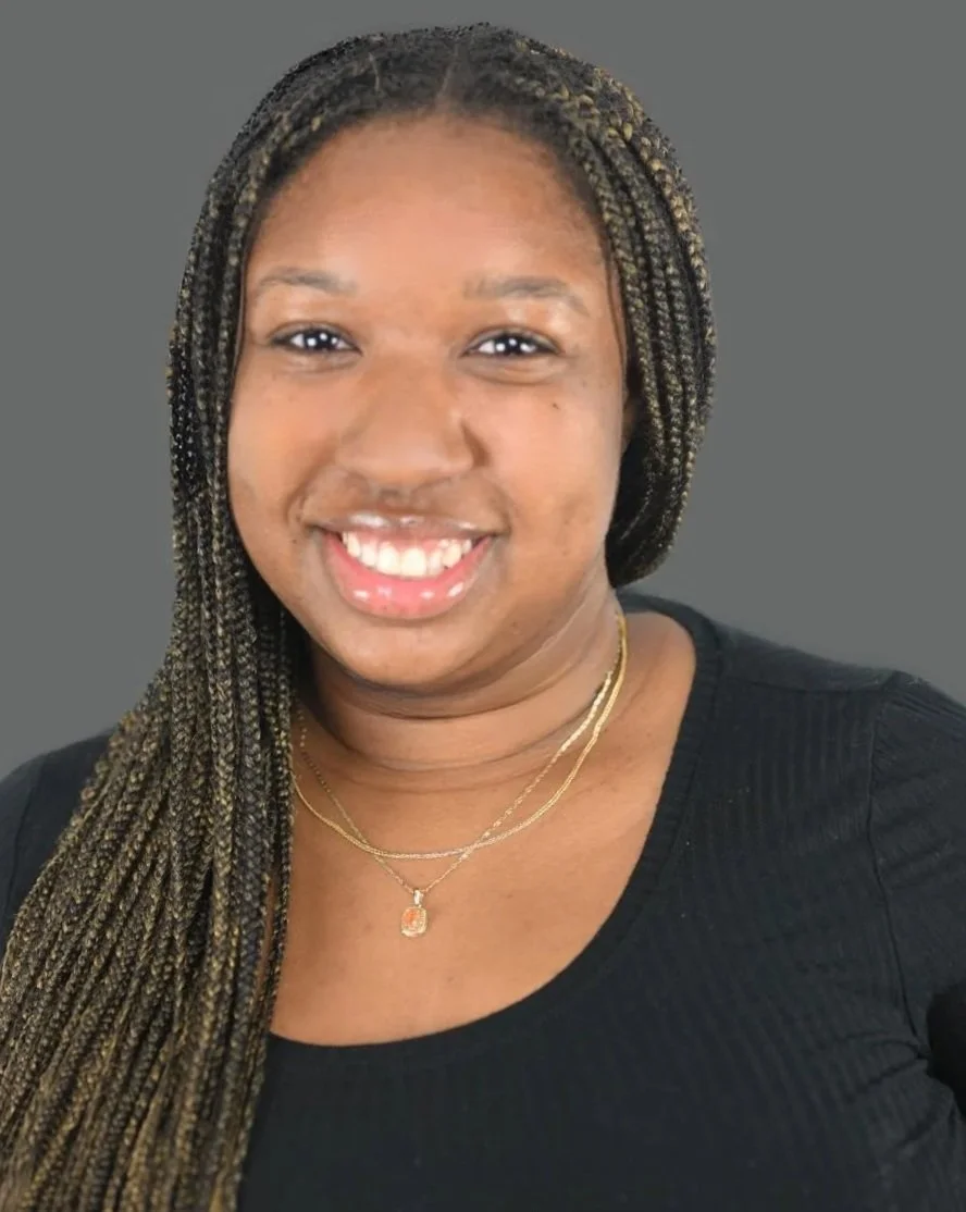A woman with long braided hair, smiling, wearing a black top and layered gold necklaces, against a gray background.