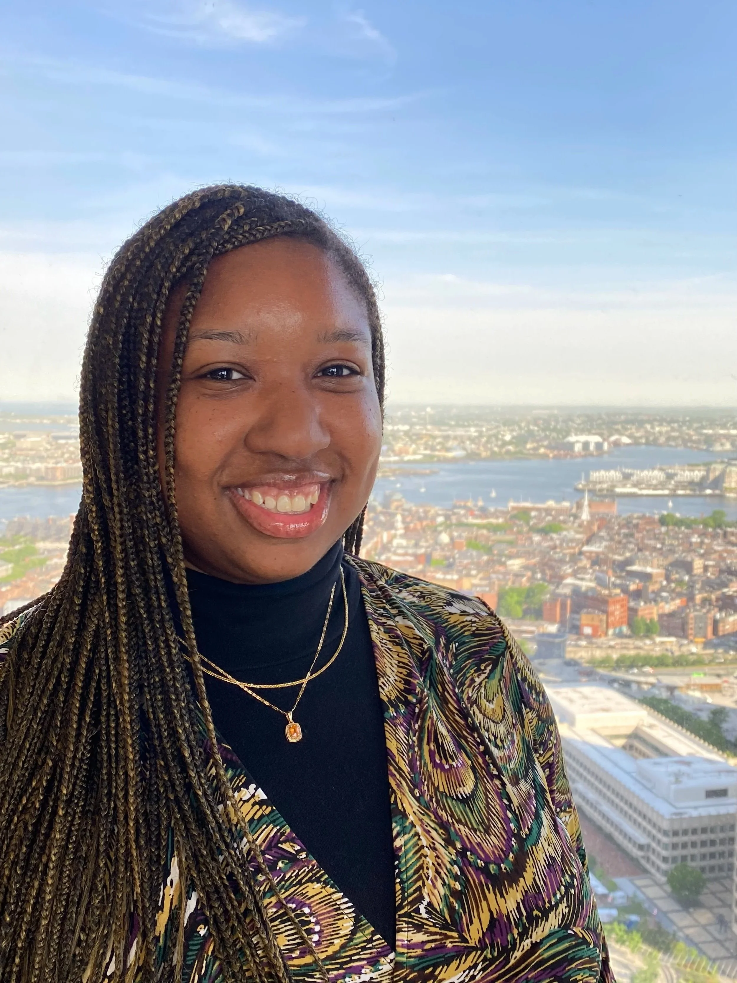 A woman with braided hair and jewelry smiling in front of a cityscape view with water and buildings under a blue sky.