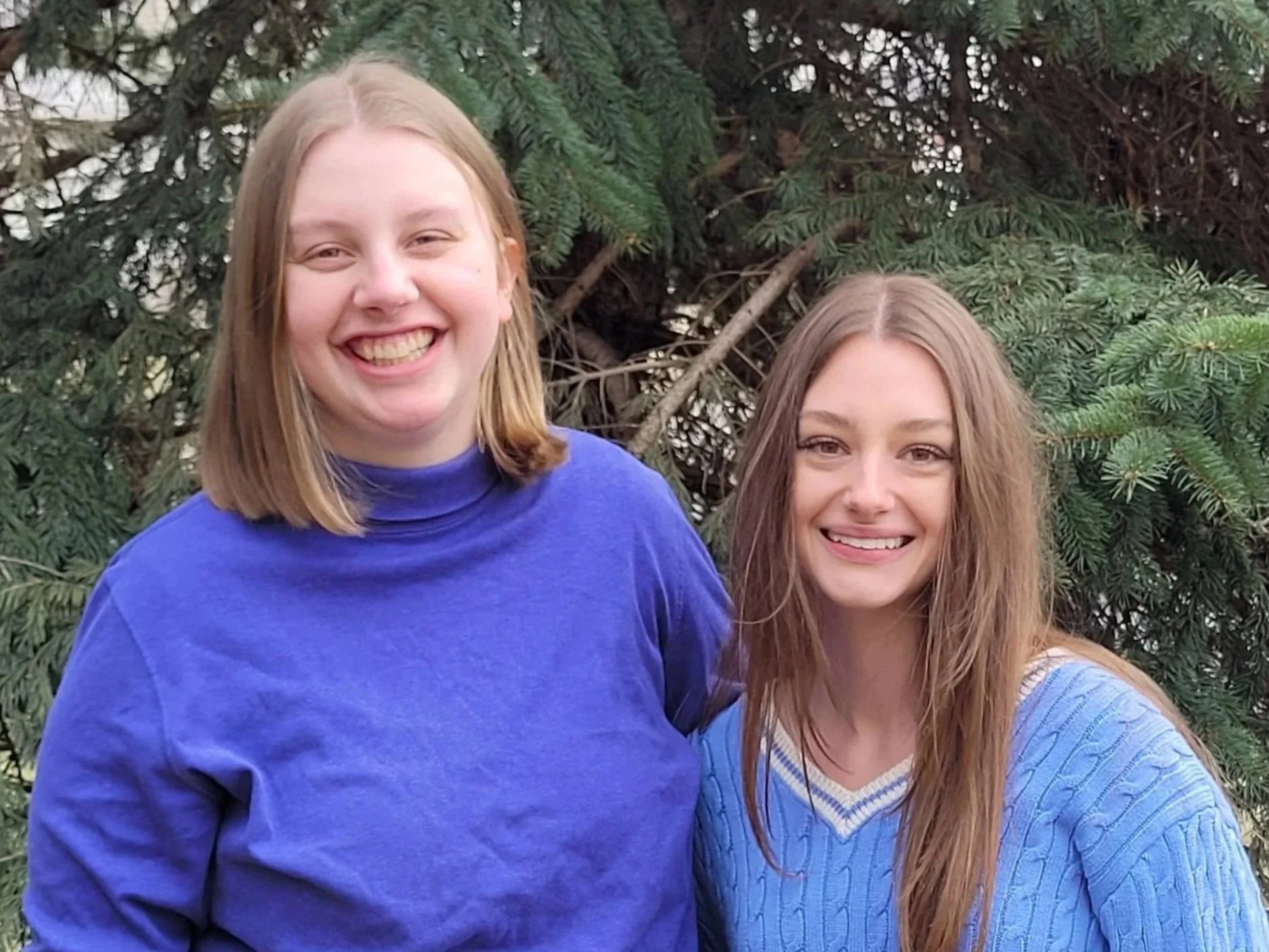 Johannah (left) has shoulder blonde hair and is wearing a periwinkle, mock-neck shirt. Lexie (Right) has long brown hair and is wearing a blue, cable-knit sweater. Both women are smiling with evergreen trees in the background.
