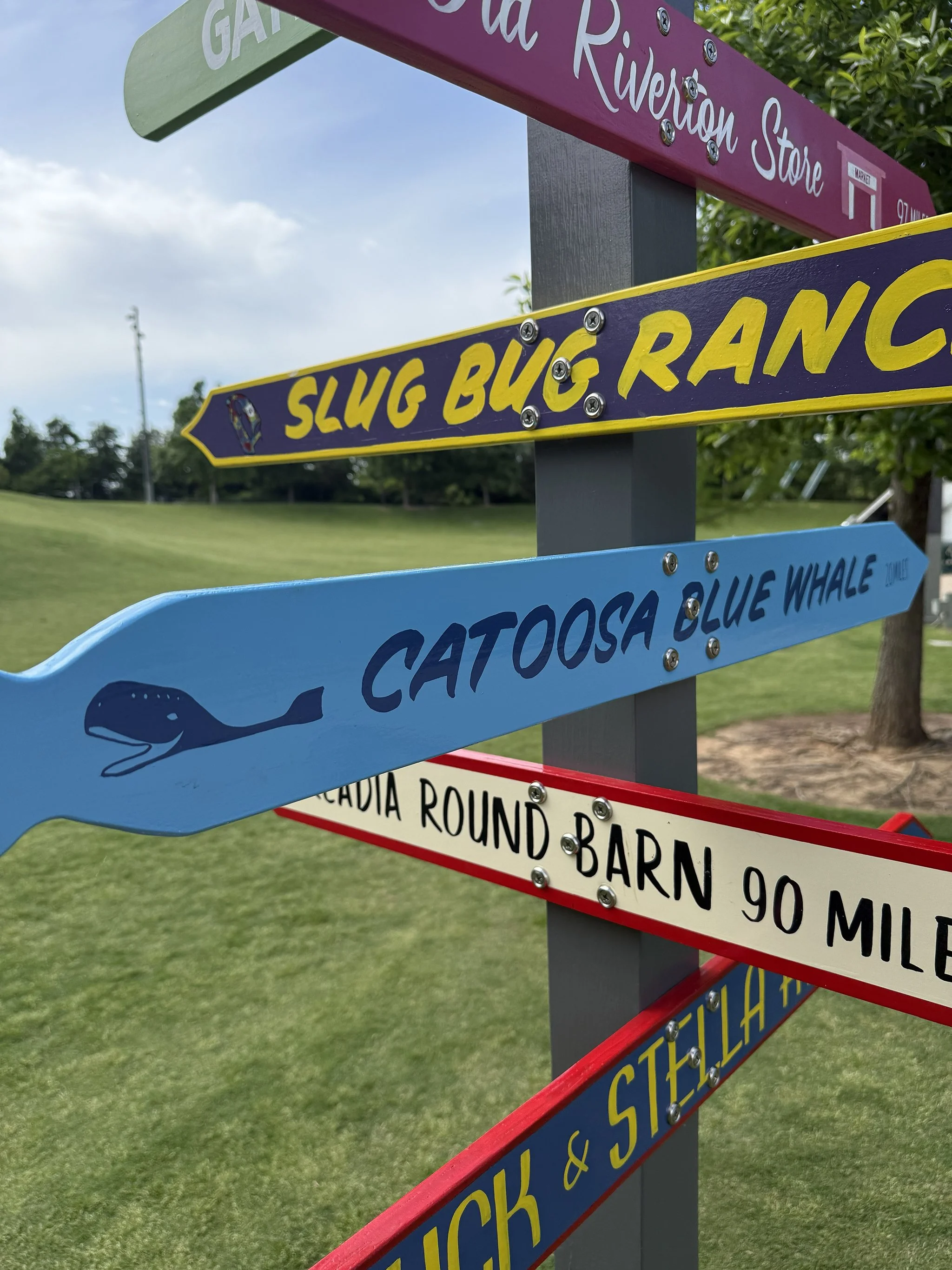 Colorful directional signs pointing to different locations, including 'Slug Bug Ranch,' 'Catoosa Blue Whale,' and others, set outdoors with grass and trees in the background.
