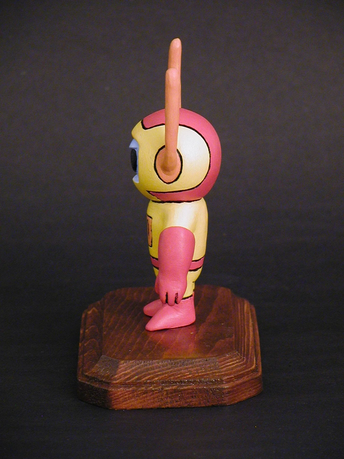 Side view of a colorful cartoon character figurine with a pink nose, yellow body, and pink limbs, standing on a wooden platform against a dark background.