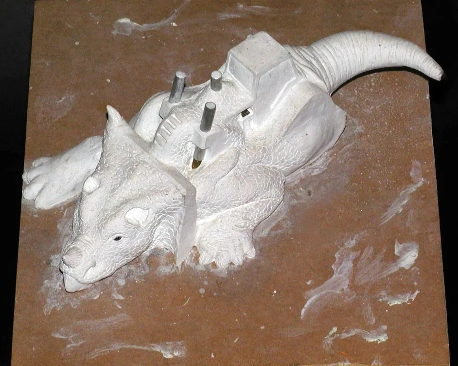A white sculpture of a dragon with detailed scales, horns, and a tail, resting on a brown work surface with some clay residue.