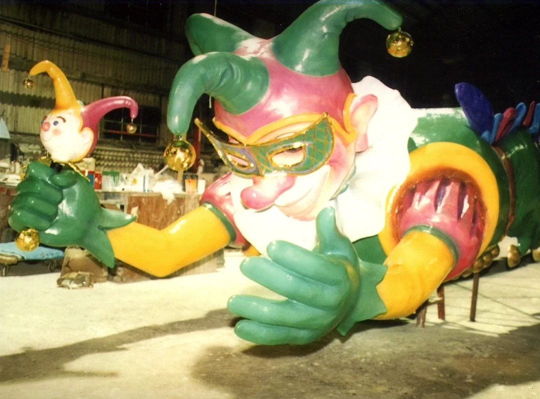 Colorful jester sculpture with a playful expression, wearing a mask, holding a small jester head, against an industrial background