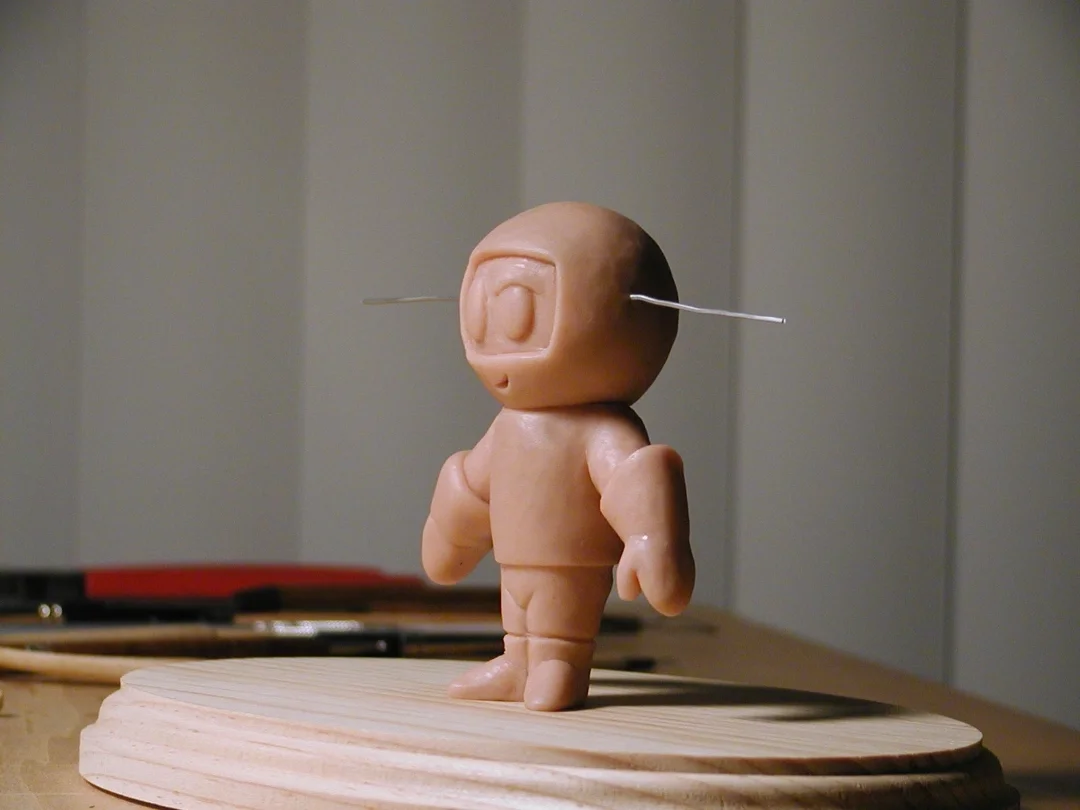 A small humanoid sculpture with a round head and an antenna, standing on a wooden base.