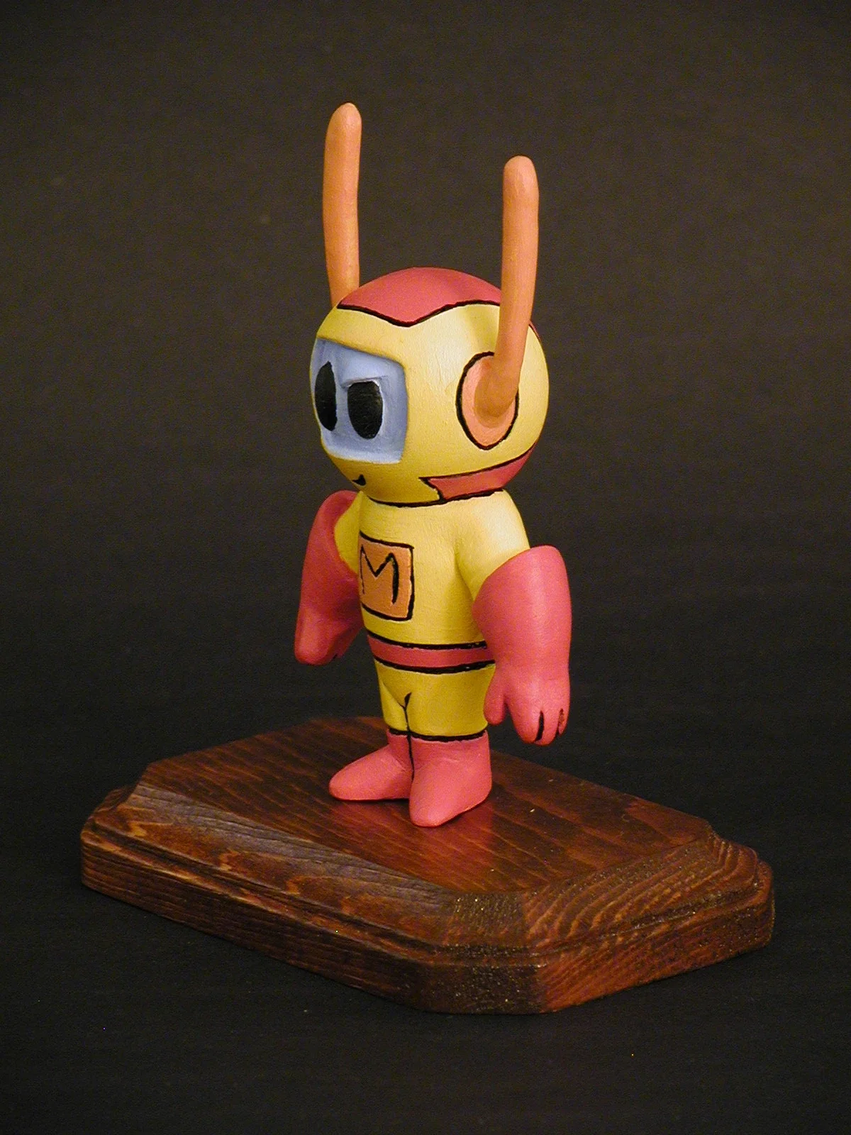 Painted figurine of a superhero character with yellow and pink costume, large eye mask, and antennae, standing on a wooden base against a dark background.