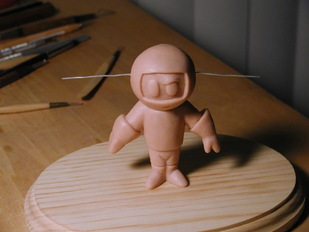 A small clay figure with a smiling face and closed eyes, standing on a round wooden platform, with a thin wire across its head, surrounded by sculpting tools on a wooden surface.