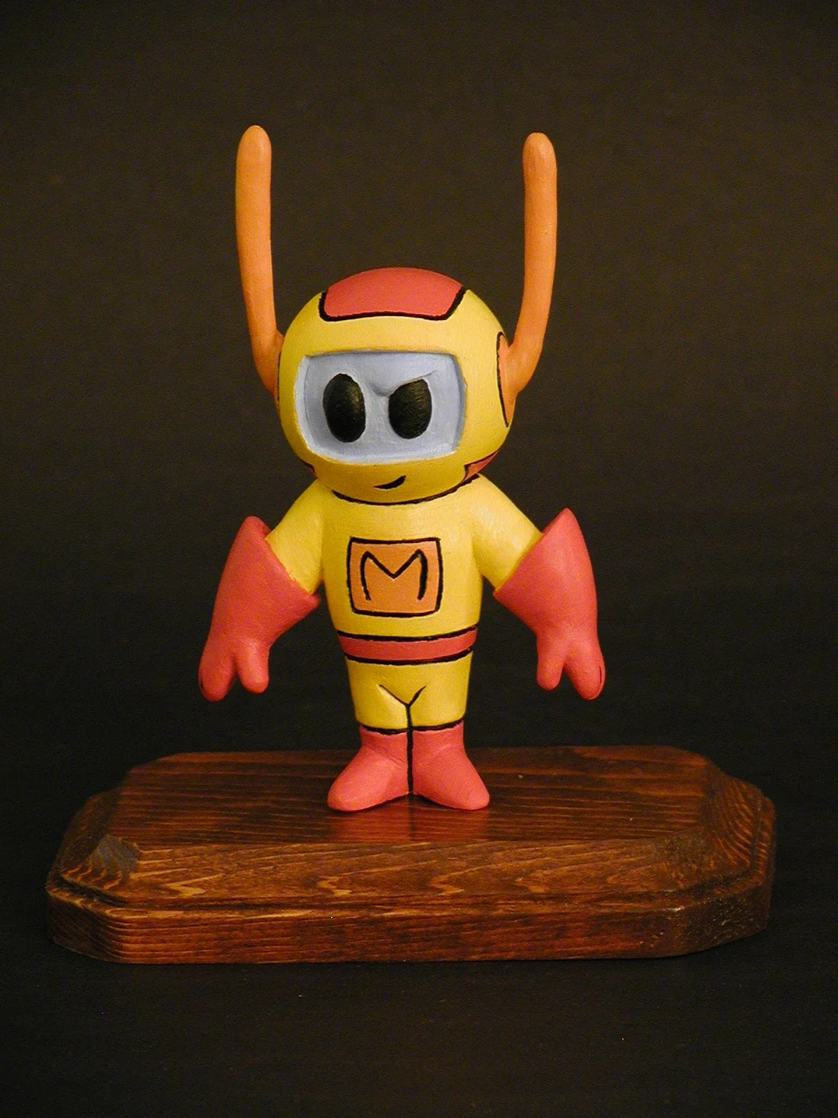 Colorful cartoon-style figure of a character with a yellow body, pink gloves, and tall pink antennae, standing on a wooden base against a dark background.