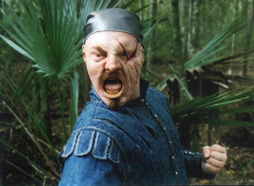A man with a torn face mask, showing a scar and open mouth, fighting aggressively in a jungle setting.
