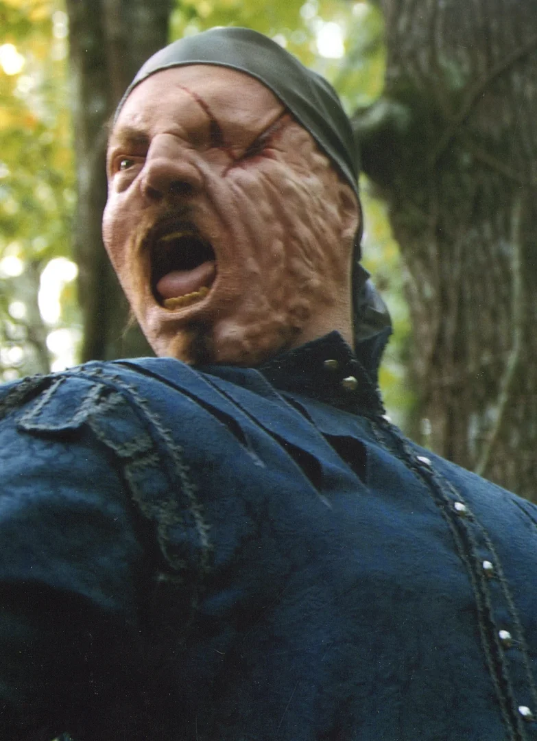 A person with a distorted, zombie-like face, wearing a black bandana and a dark denim jacket, mid-shout in a wooded area.